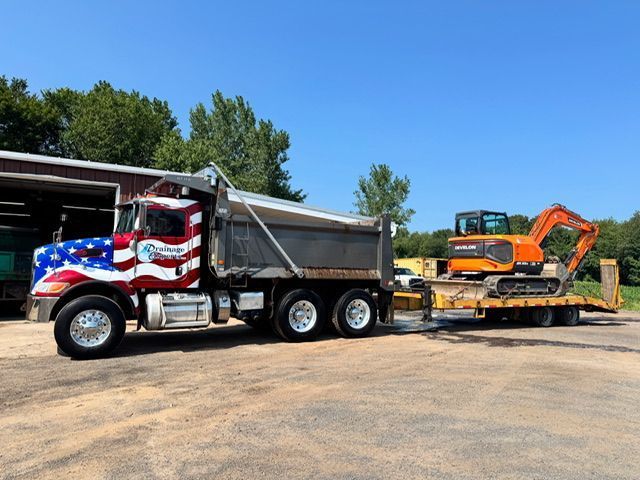 American flag-painted dump truck towing an orange excavator on a trailer, parked on a sunny day.