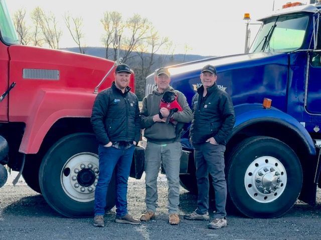 Three men standing in front of two semi-trucks; one red, one blue. A man holds a small dog.