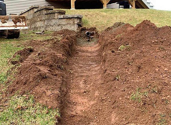 A trench dug in a yard, leading towards a retaining wall. Two pipes are visible within the trench.