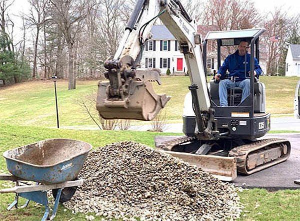 Man operating a small excavator, moving gravel next to a wheelbarrow in a yard.