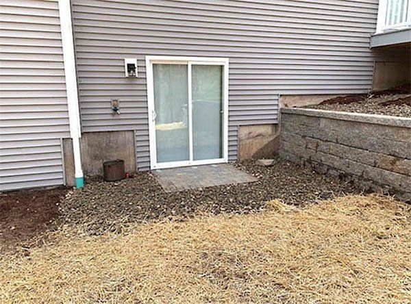 Sliding glass door at the base of a building, surrounded by gravel and a retaining wall.