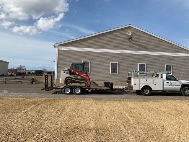 A white truck towing a trailer with a Bobcat in front of a light gray building.