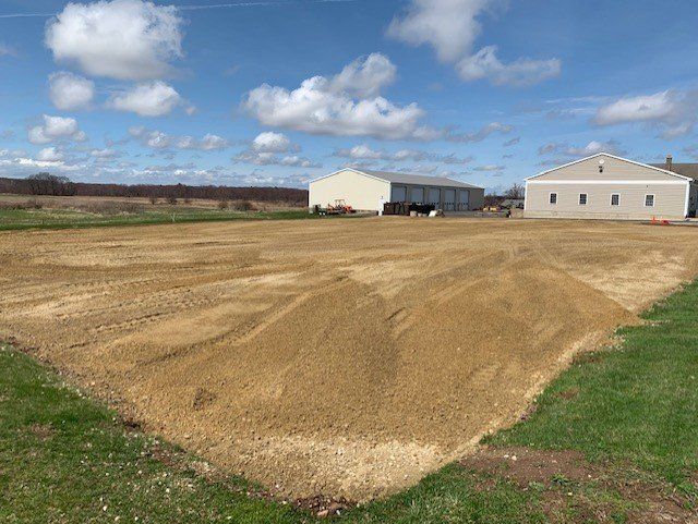 A leveled dirt pad in a field with two buildings and a blue sky with clouds.