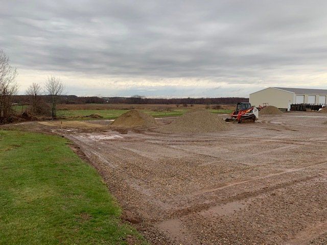 Gravel and dirt piles next to a skid steer in a muddy construction site near a building and grass. Overcast sky.