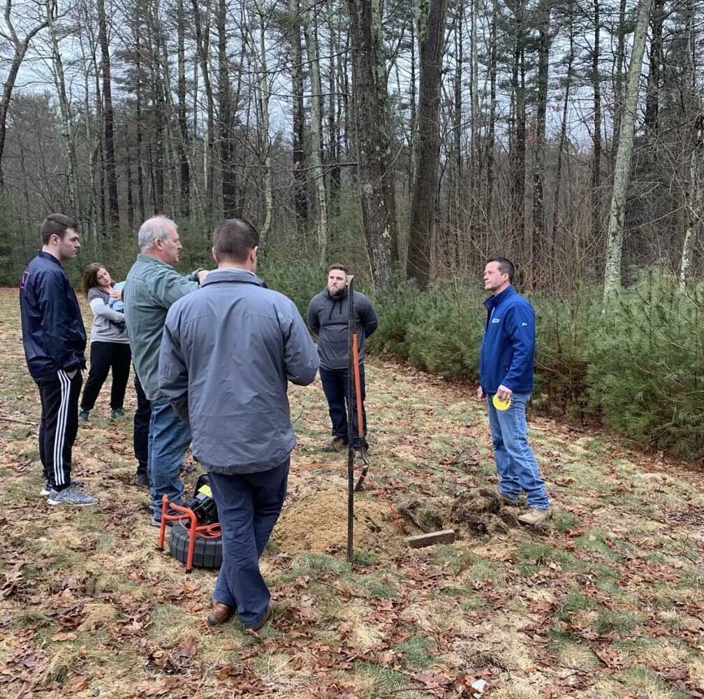 Group of people outdoors observing a dug hole. Several are gesturing, some are holding tools, and the area is wooded.