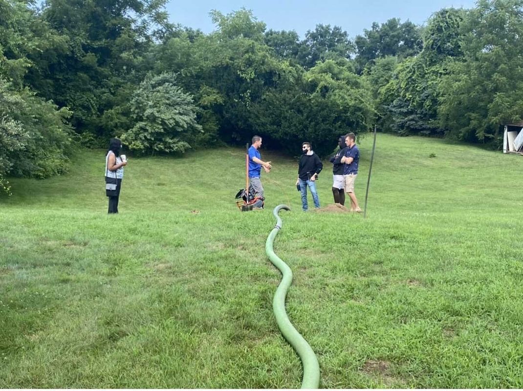 People gather on a grassy hill; a green hose runs toward the group. Trees in the background.