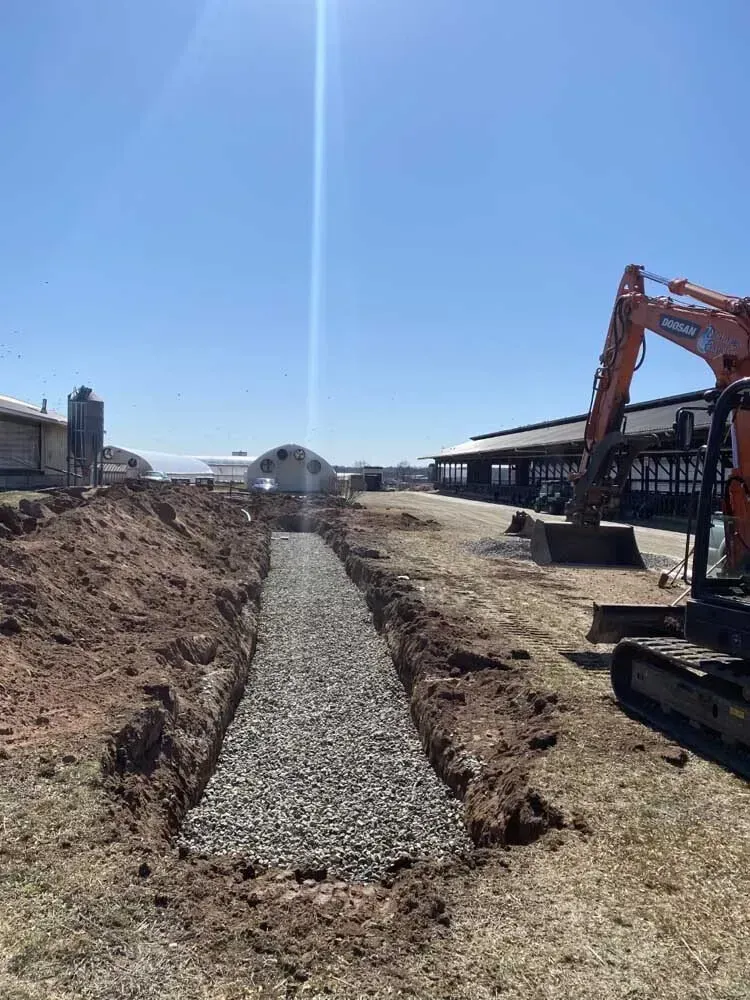 An excavator digs near a trench filled with gravel on a farm, sunny day.