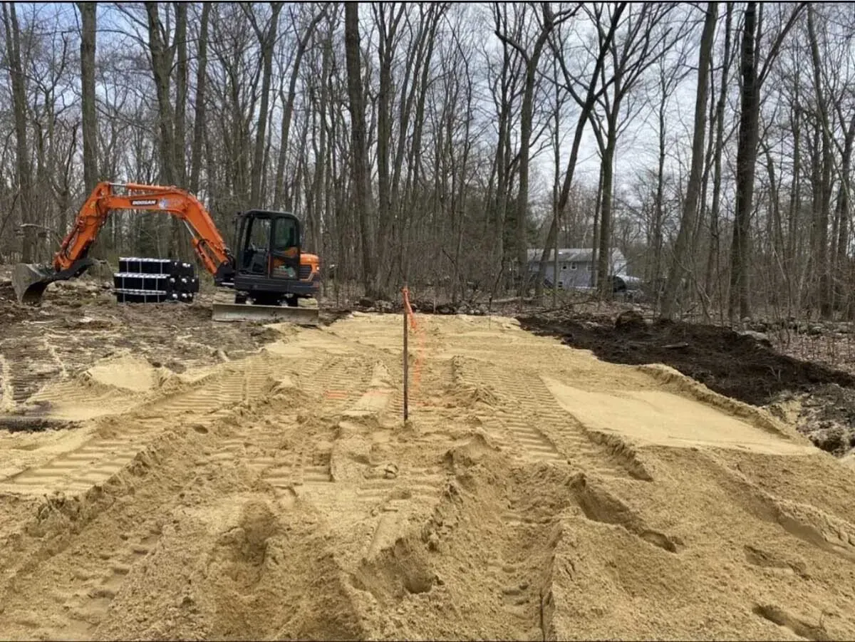 Construction site with an excavator and a large pile of sand in a wooded area.