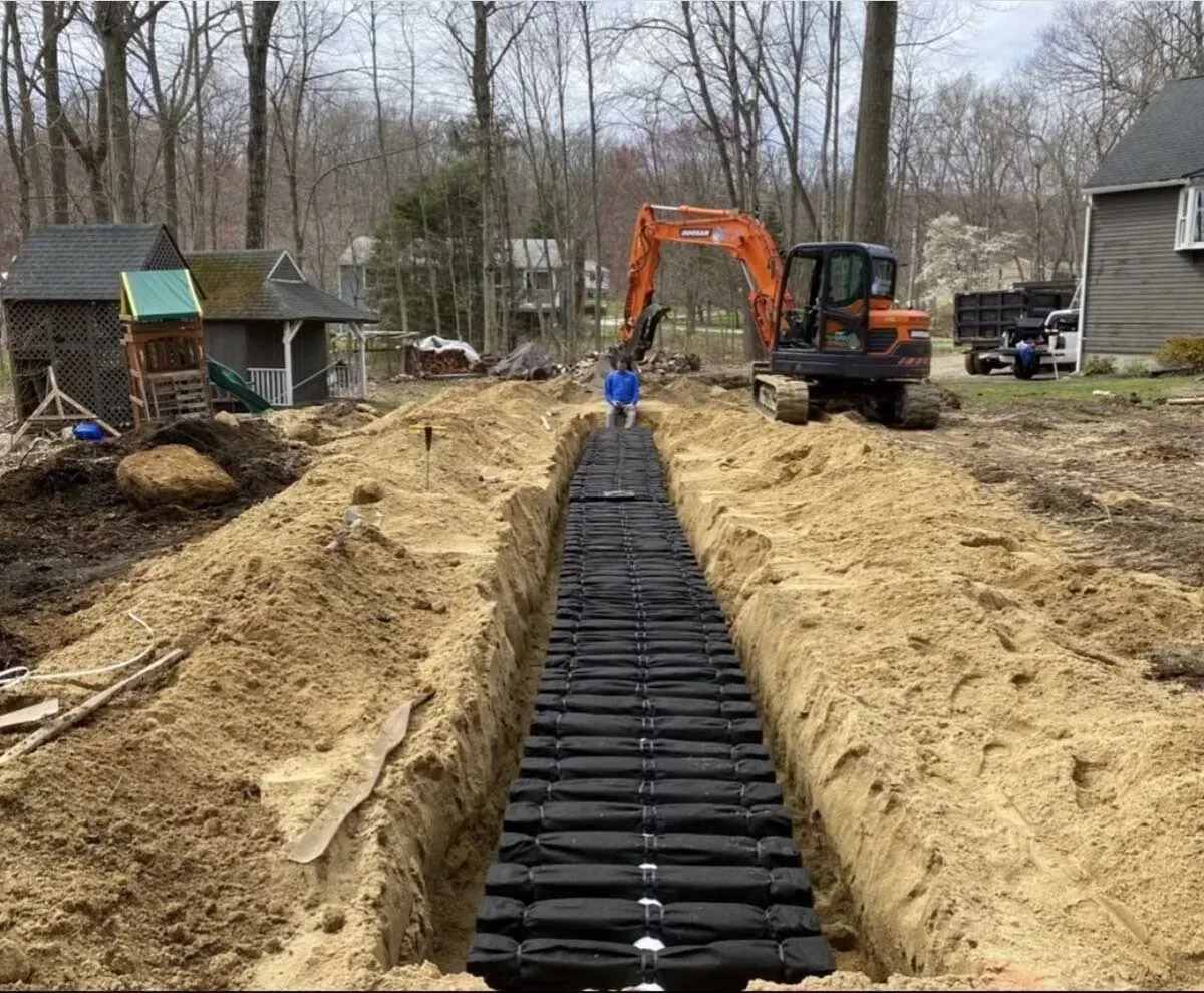 Construction site with long trench, black drainage system, excavator, and person.