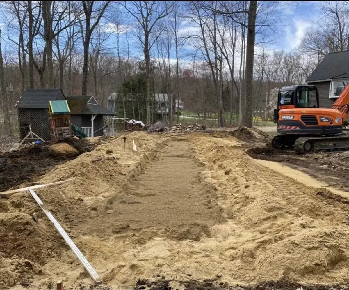 Excavated ground for a construction project, orange excavator on right. House and trees in the background.