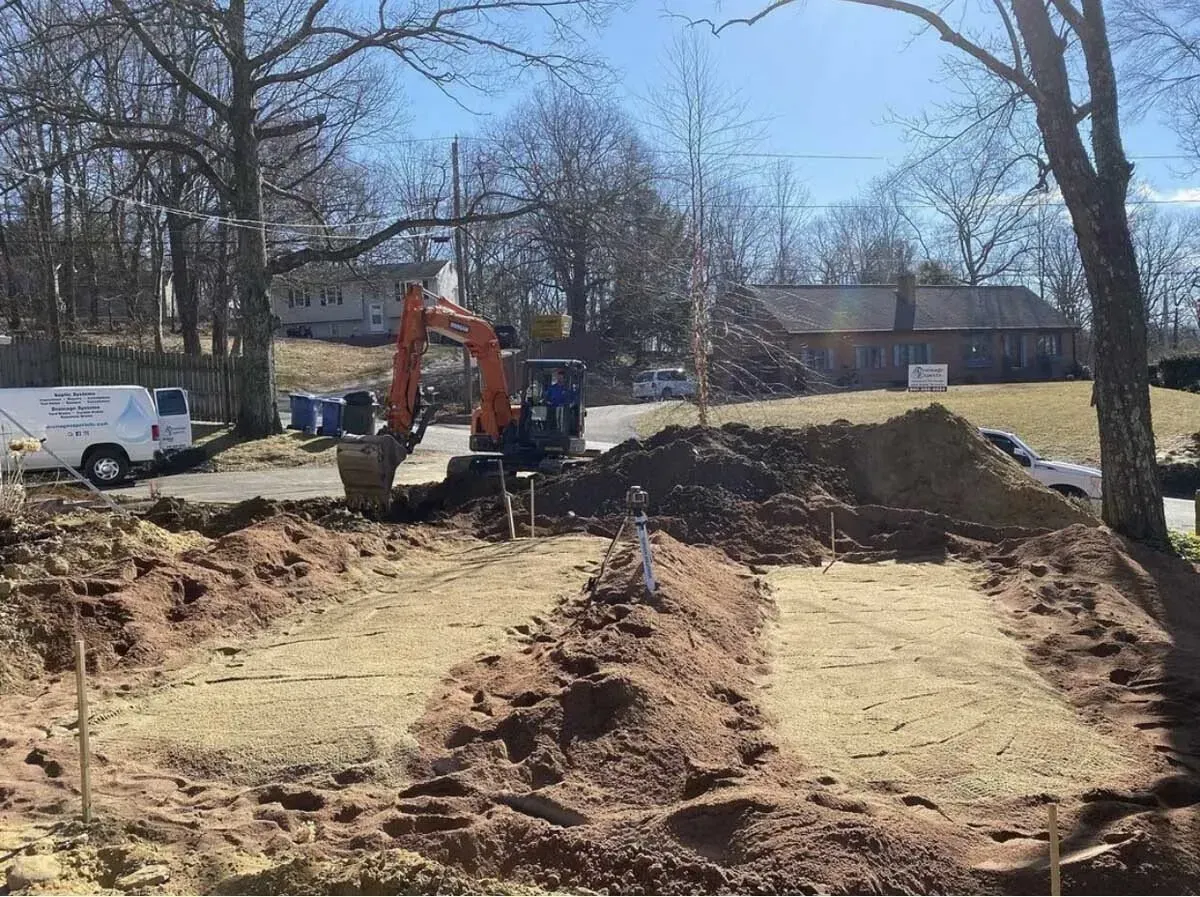 Excavator digging a foundation in a residential area; dirt piles, blue sky.