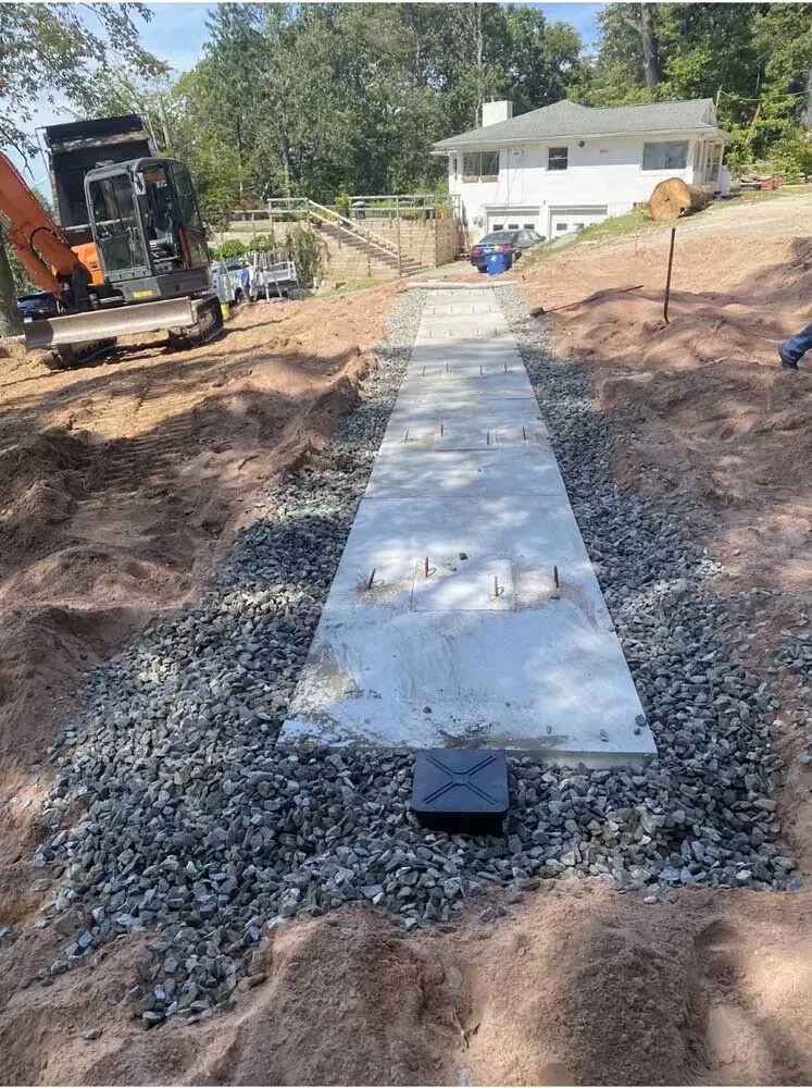 Construction site with concrete pathway, gravel, and heavy machinery, leading to a white house on a hill.