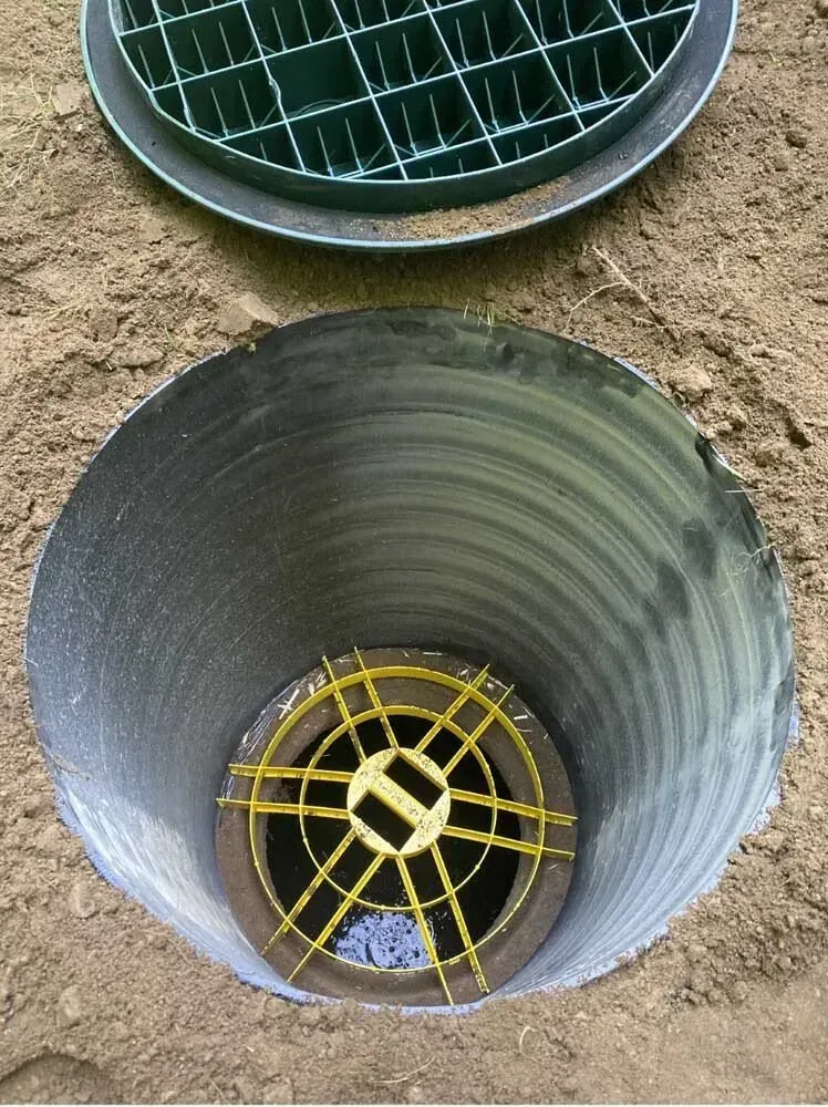 Open, corrugated pipe in the ground with a grate at the bottom and a matching lid.