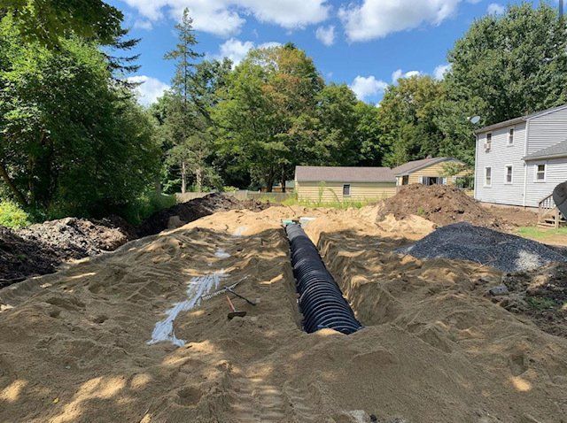 Black drainage pipe in a sandy trench; construction site with trees and buildings in the background.