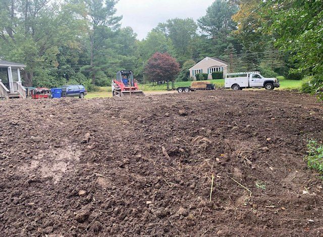 Dirt work in progress; red and blue machinery on a freshly tilled yard, a white truck, and a house in the background.