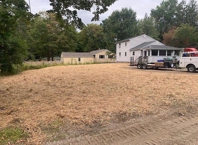 Cleared lot covered in wood chips with two buildings and work trucks in the background.