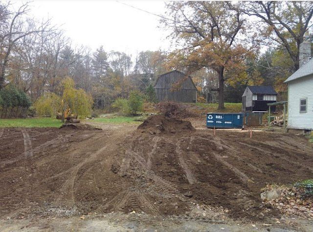 Dirt piles and tractor tracks on a cleared lot with a barn and house in the background.