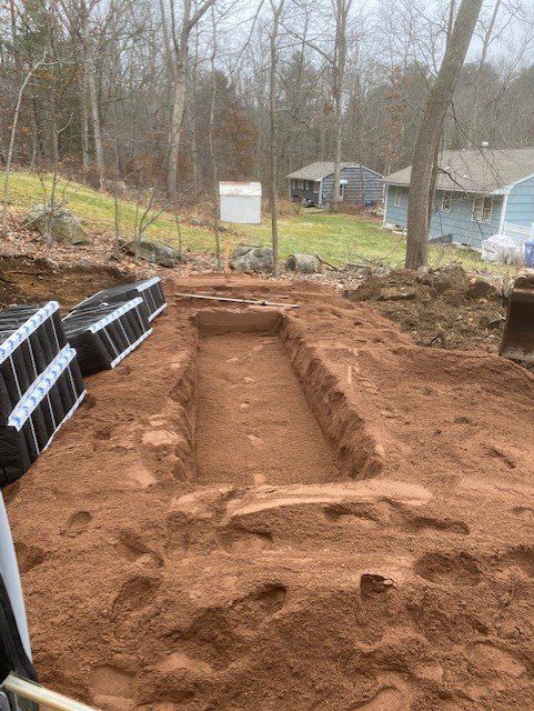Excavated rectangular pit in red soil, with landscaping blocks at left, trees, and houses in background.