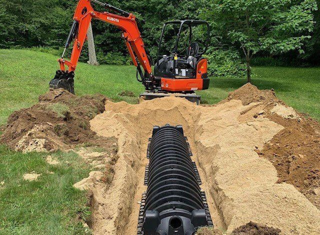 Orange excavator digging trench, next to black septic system, on a lawn.
