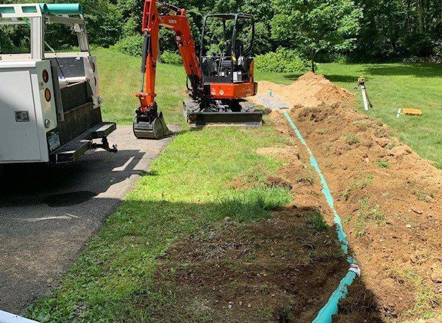 An orange excavator and truck working on a green pipe in a trench, outdoors on a sunny day.
