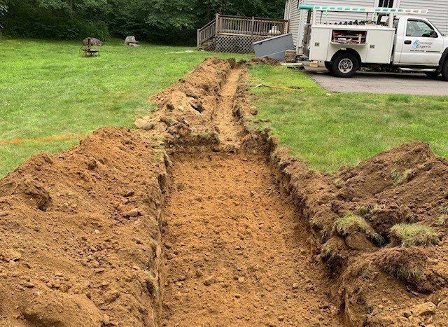 A long trench dug in a grassy yard, with a work truck parked nearby.