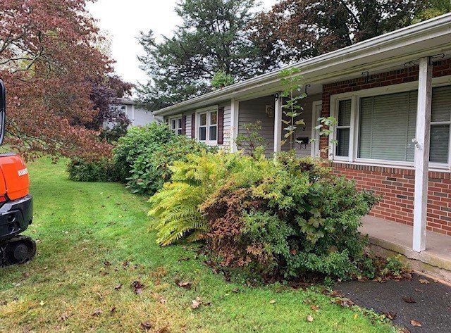 A house with overgrown bushes and a mini excavator on the lawn. Green grass and brick facade.