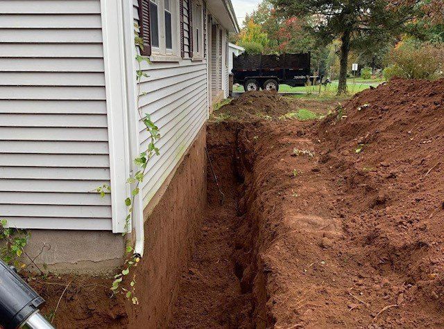A trench dug alongside a house, ready for foundation work. Red-brown soil, beige siding.