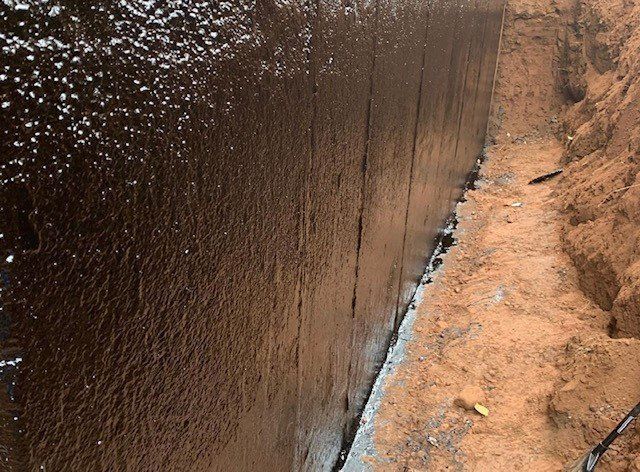 Black waterproofing coating on a concrete foundation wall in an excavated trench.