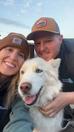 Couple smiling with a white Husky dog; all outdoors, wearing hats and jackets, under a blue sky.
