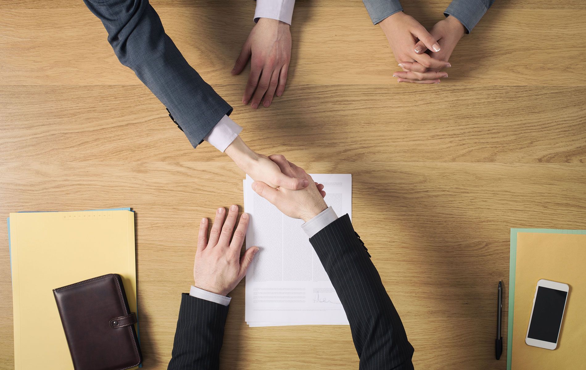 Two people in suits shaking hands over a contract on a wooden table.