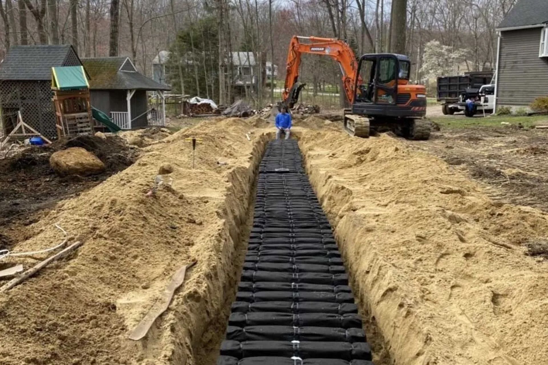 A long trench with a black drainage system, an orange excavator, and a man standing in the trench.