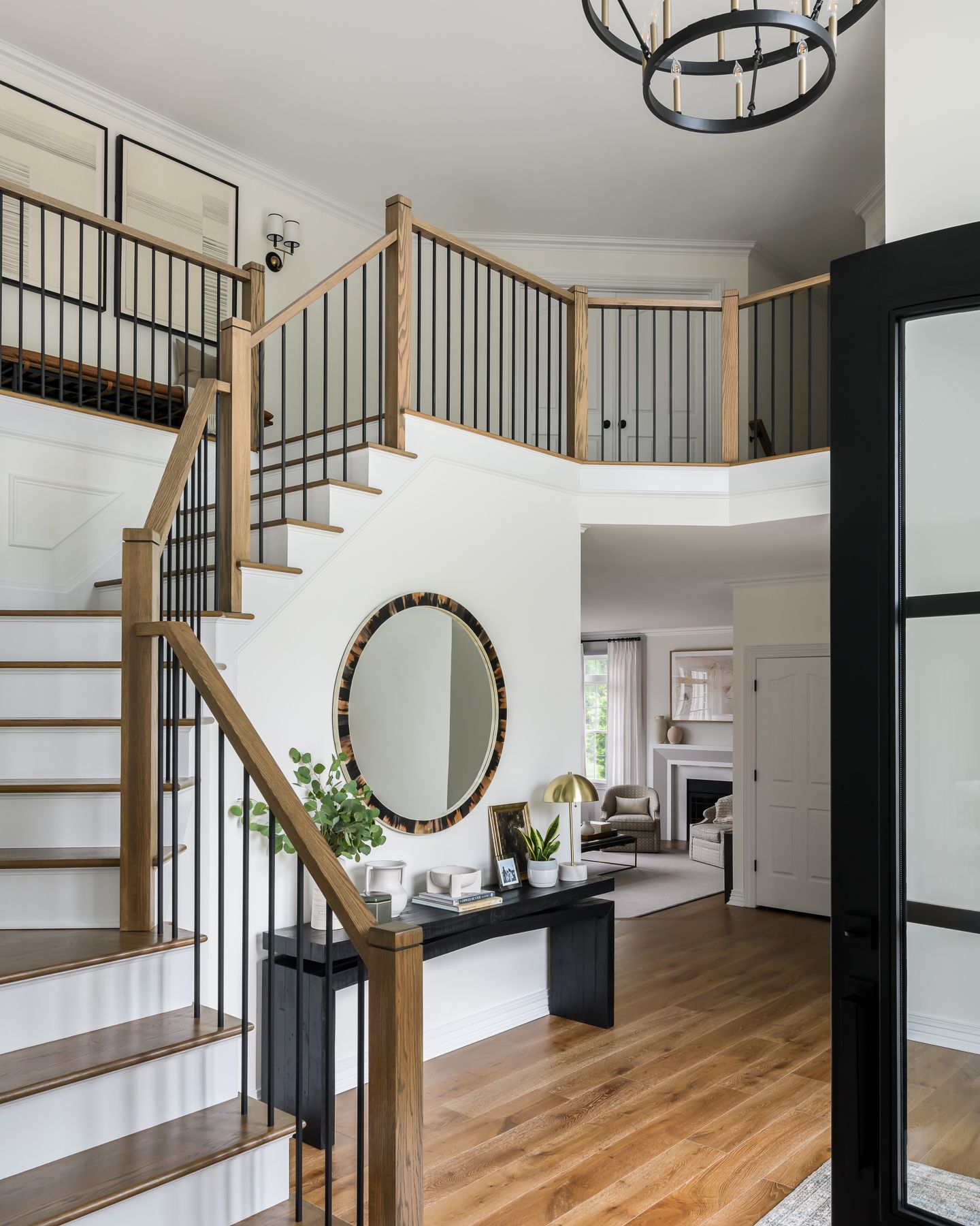 a bright entry hall with wooden staircase, black metal railings, and a large round mirror