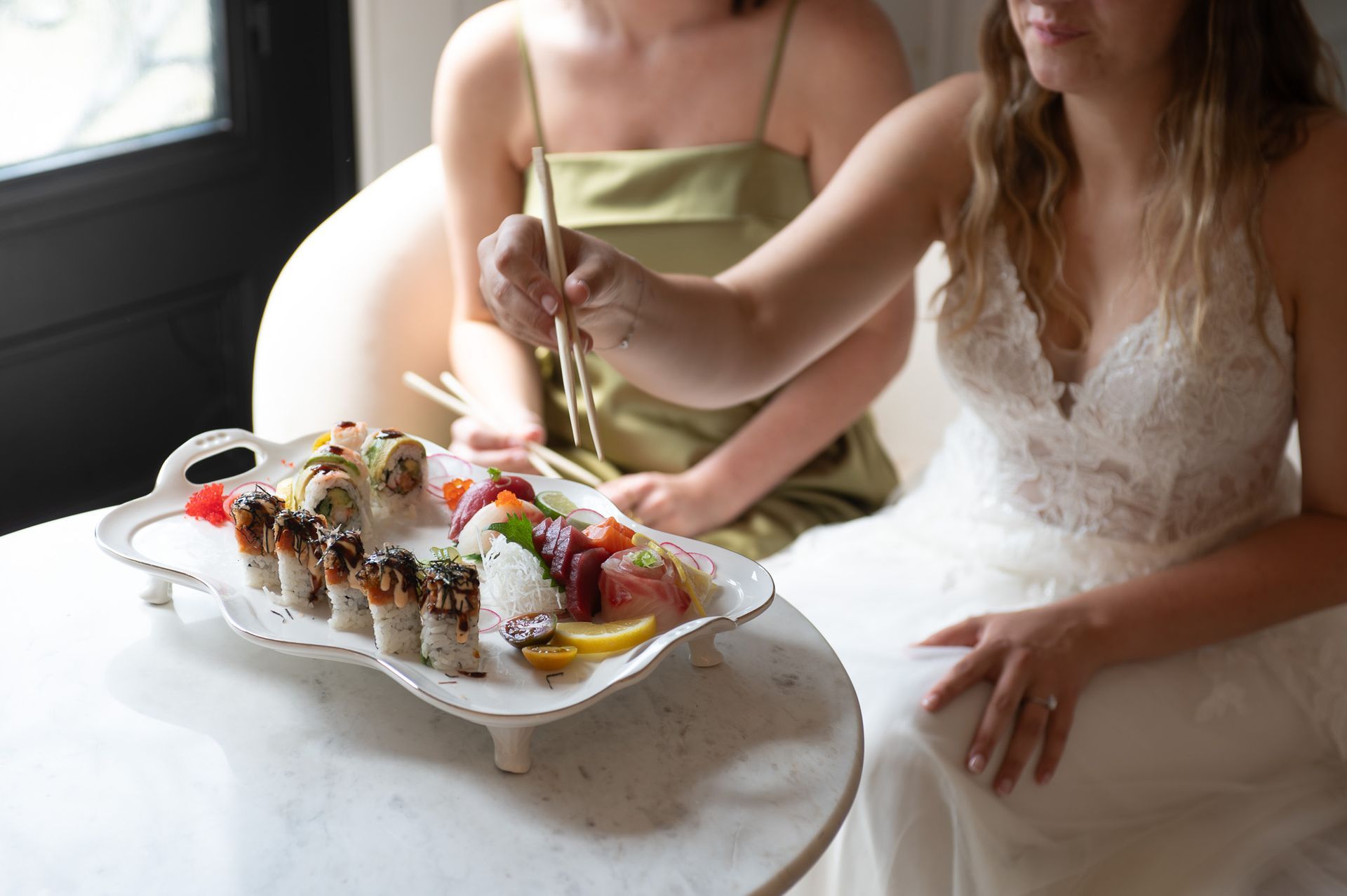 Two people at a table sharing sushi, one in a white dress reaching with chopsticks.