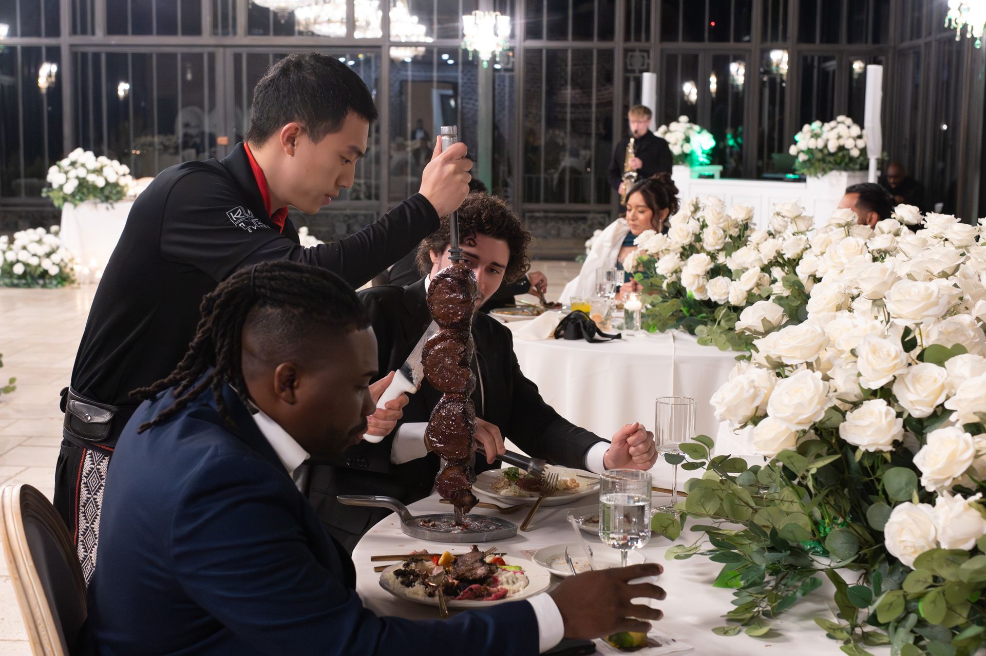 Guests dine at a floral banquet table while one man pours a drink amid white flower arrangements.