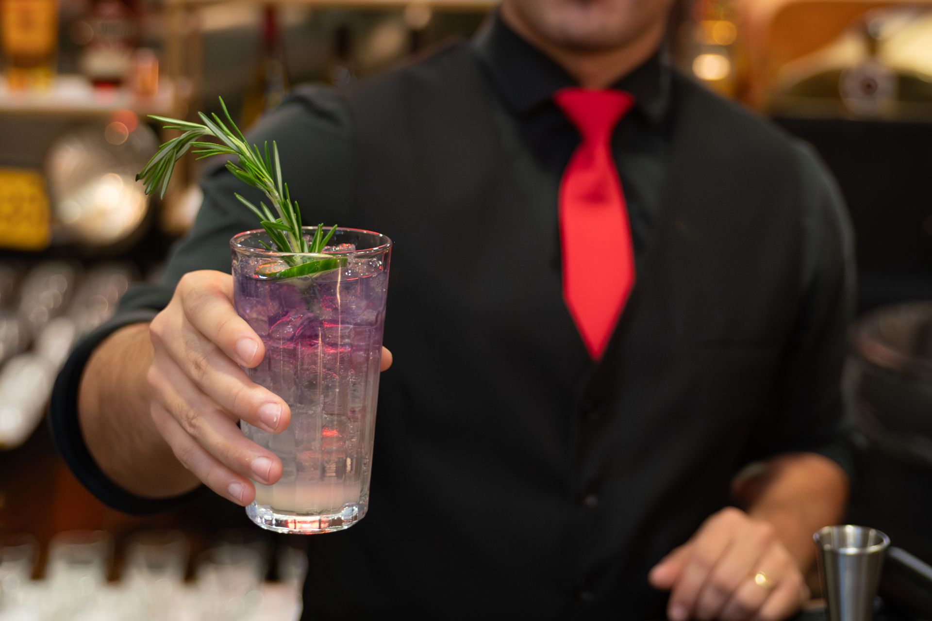 Bartender holding a purple cocktail with rosemary garnish and a red tie behind the bar