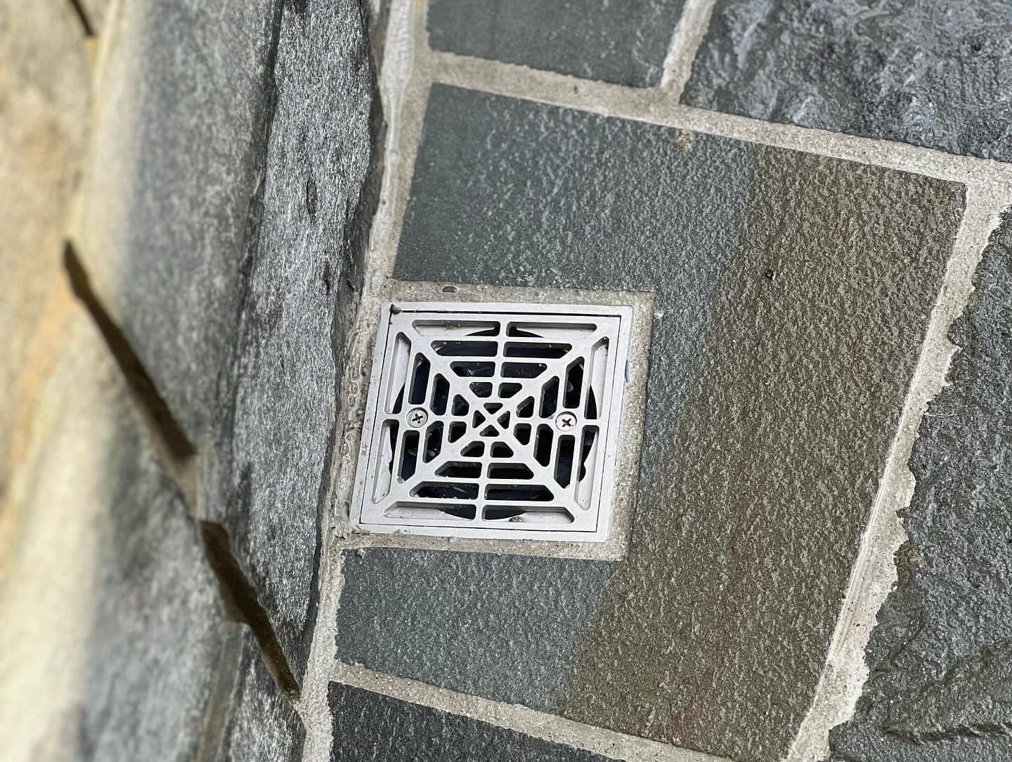a square drain on a tiled floor next to a stone wall .