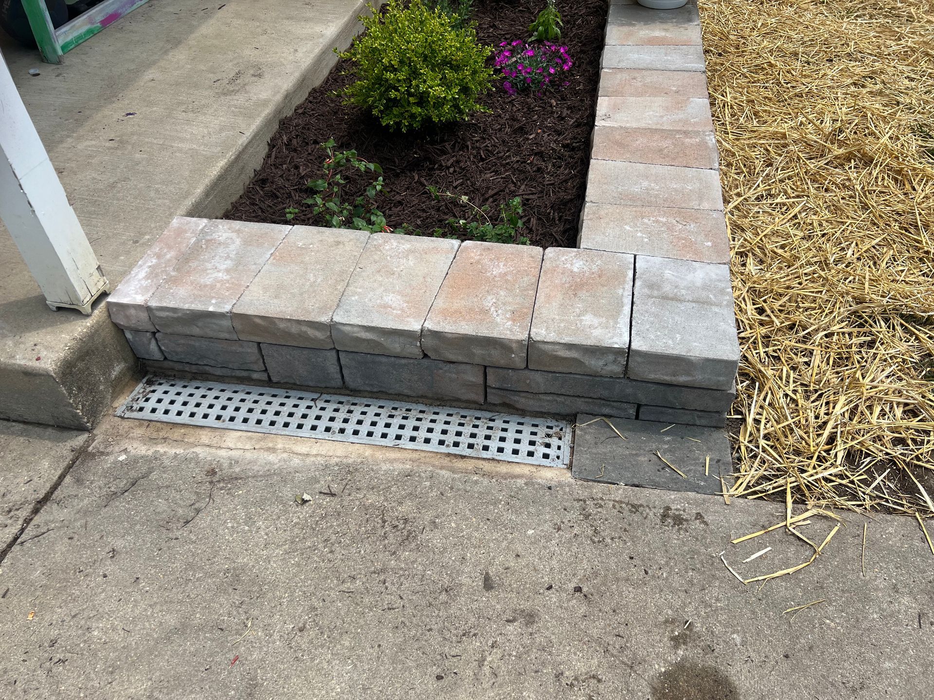 there is a drain on the sidewalk next to a garden .