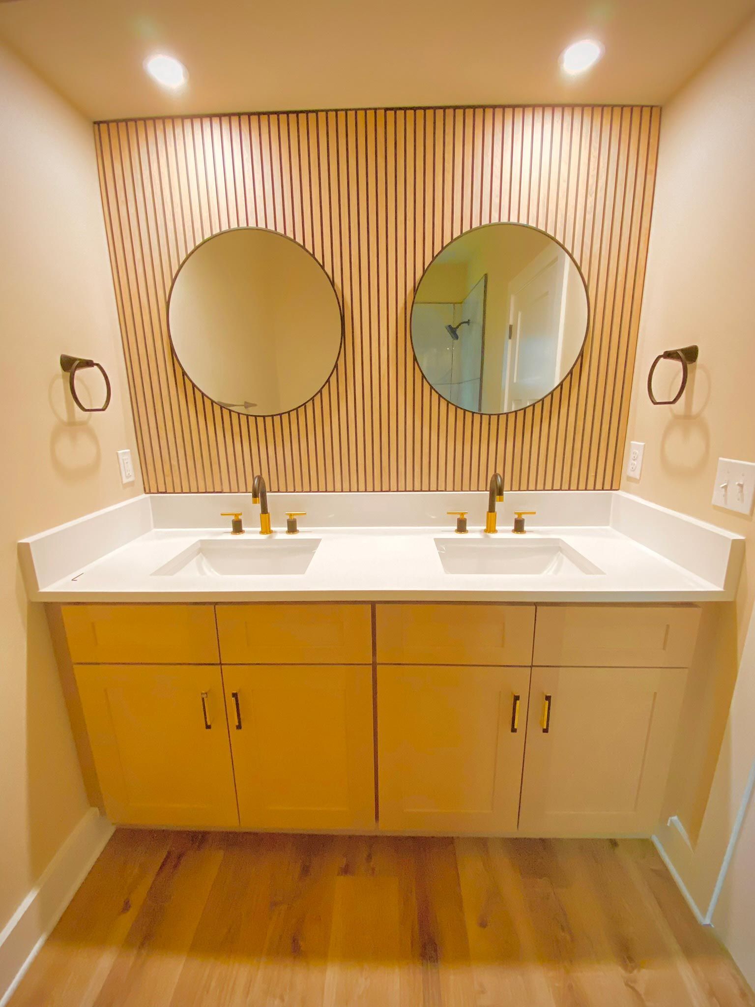 Bathroom with two sinks, round mirrors, wooden paneling, gold fixtures, and yellow cabinets.