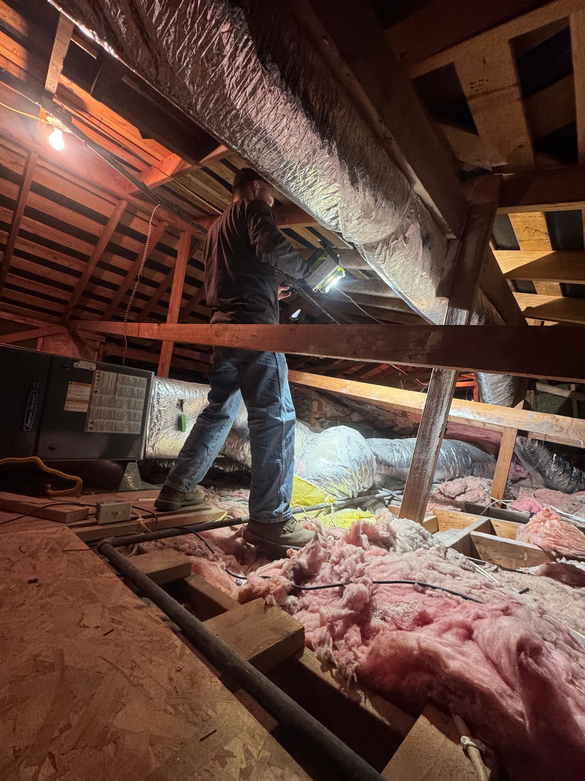 Insulated attic with exposed beams, ductwork, and pink insulation under a lit opening.