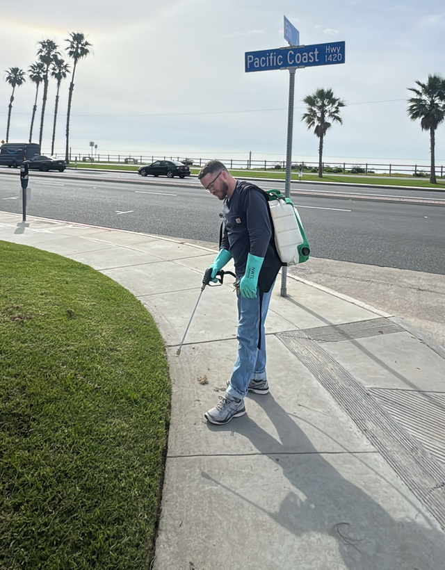 Person in teal jacket pointing at sidewalk near a street sign by the coast on a sunny day