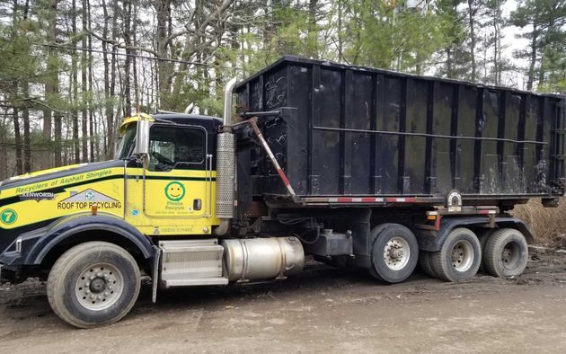 A yellow and black roll-off dump truck parked on a dirt path with trees in the background.