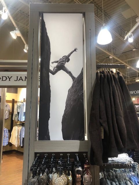 Clothing store display with a black-and-white framed artwork above hanging dark jackets and bottles below