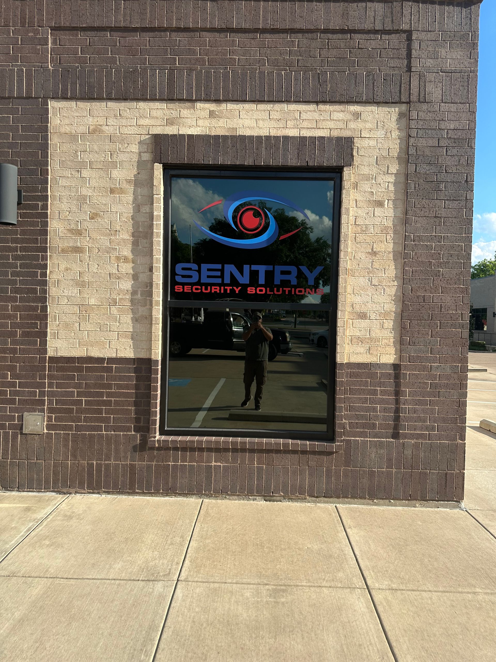 Sentry Security Solutions logo on a window in a brick building exterior, with sidewalk and sky reflected.
