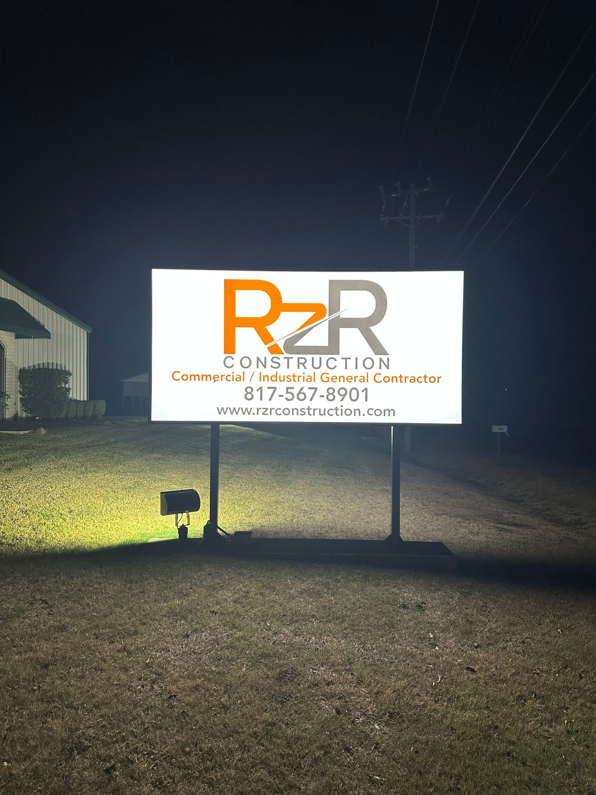 Illuminated R&R Construction sign at night on a roadside, with a building in the background