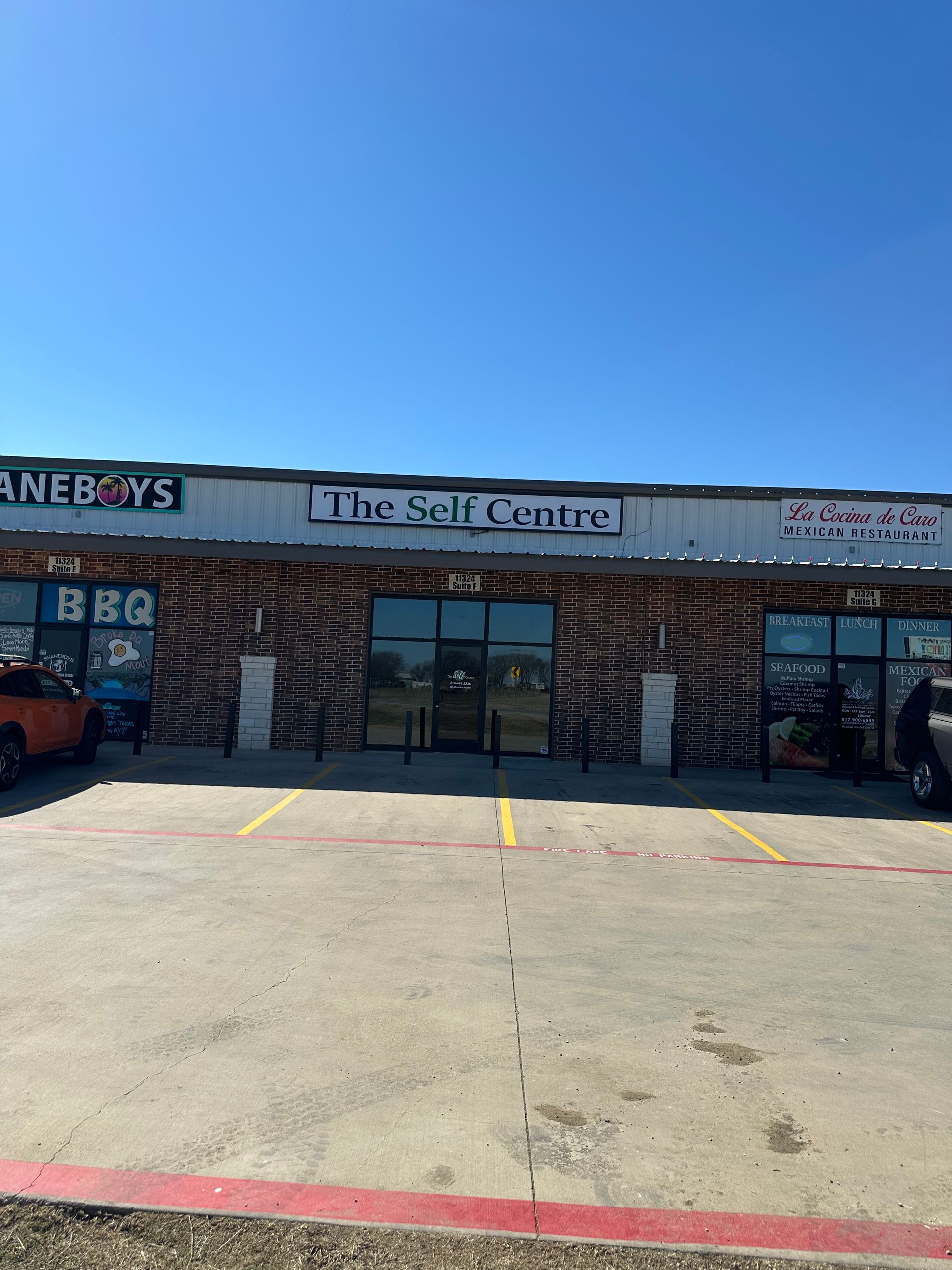 Strip mall storefront with “The Self Centre” sign, glass doors, and empty parking spaces under a blue sky
