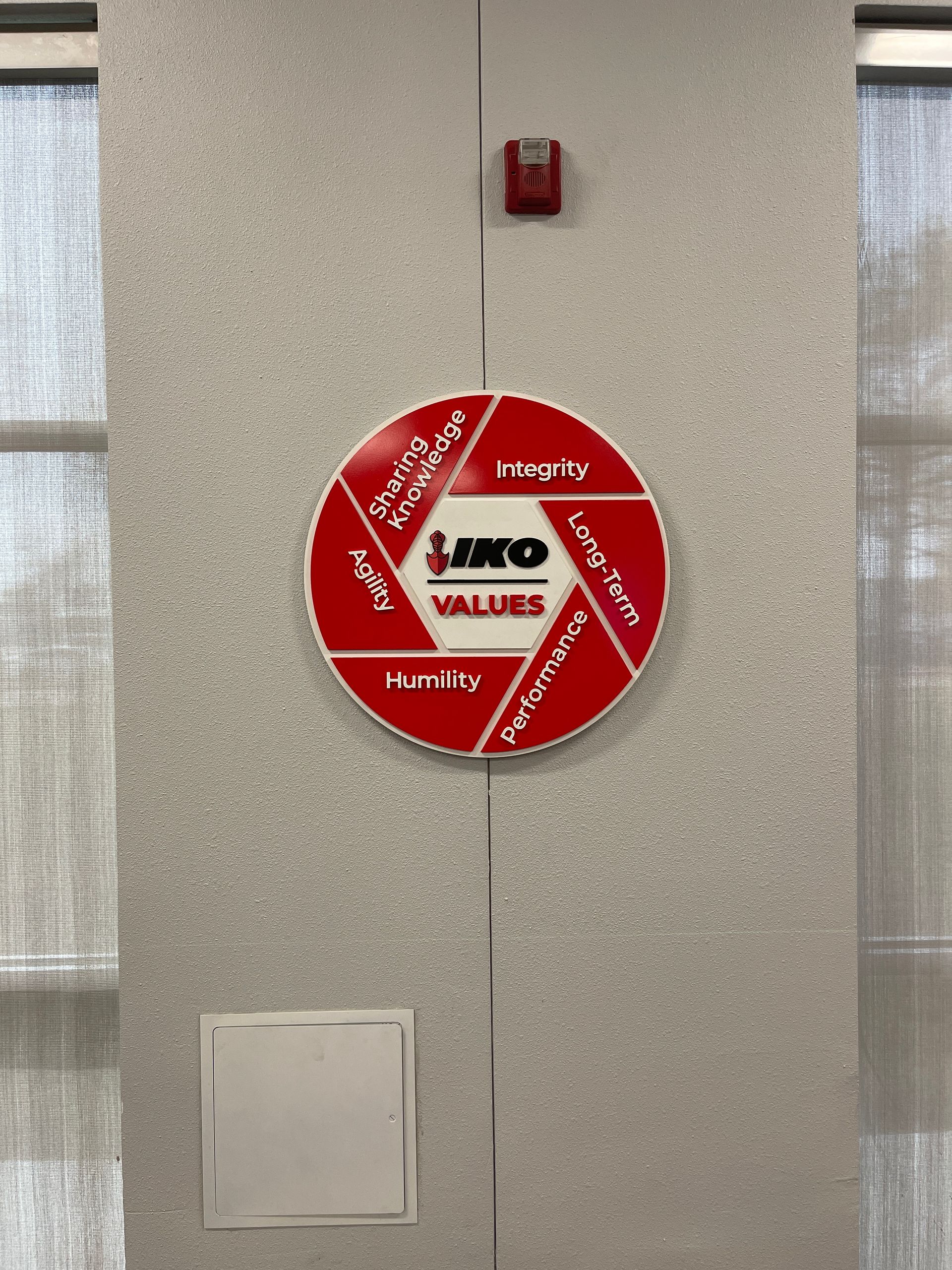 Red fire alarm bell with “Janco” label on a gray wall, above a small access panel.