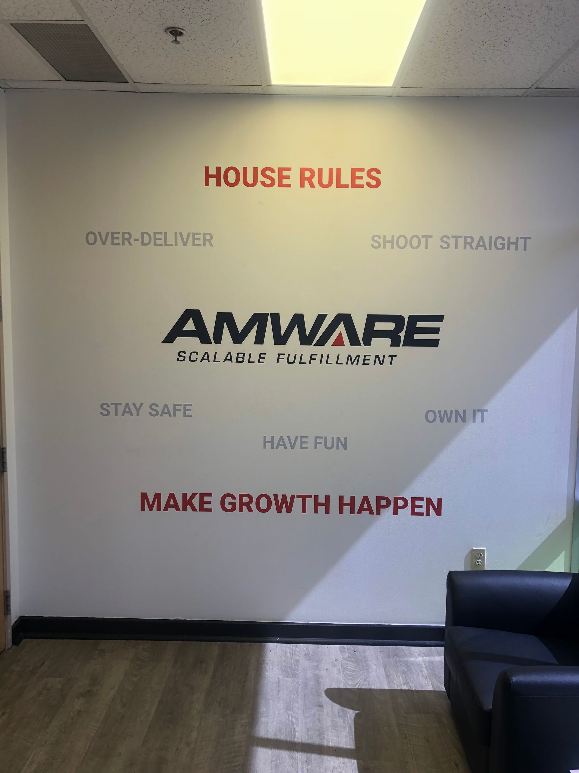 Office wall with AMWARE logo and house rules text in red and black, above a black couch and wood floor