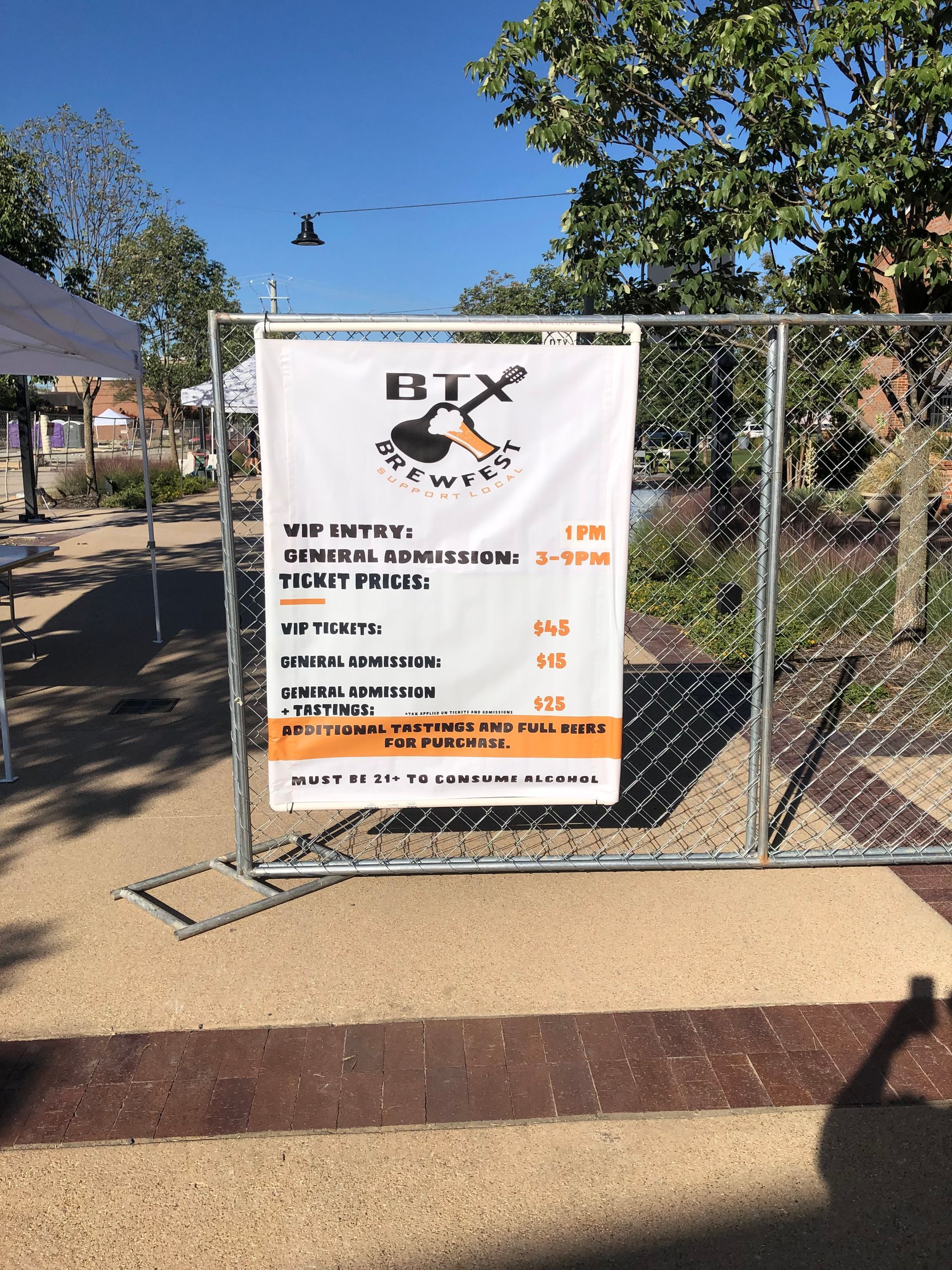 Banner on a chain-link fence at an outdoor entrance, with trees and a dirt path behind it.