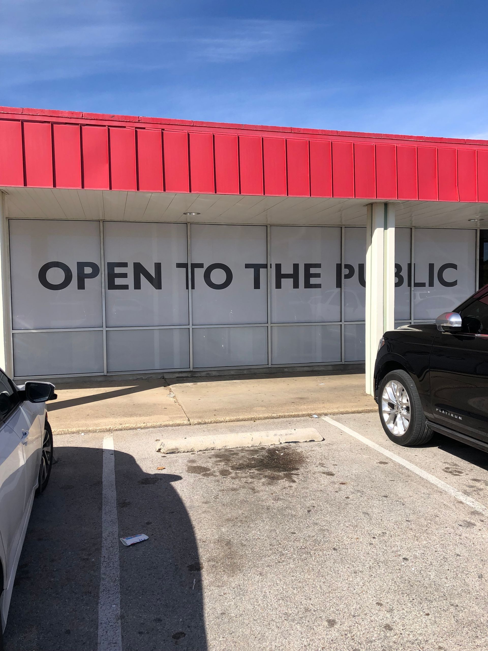 Storefront with red awning and “OPEN TO THE PUBLIC” sign, viewed from a parking lot