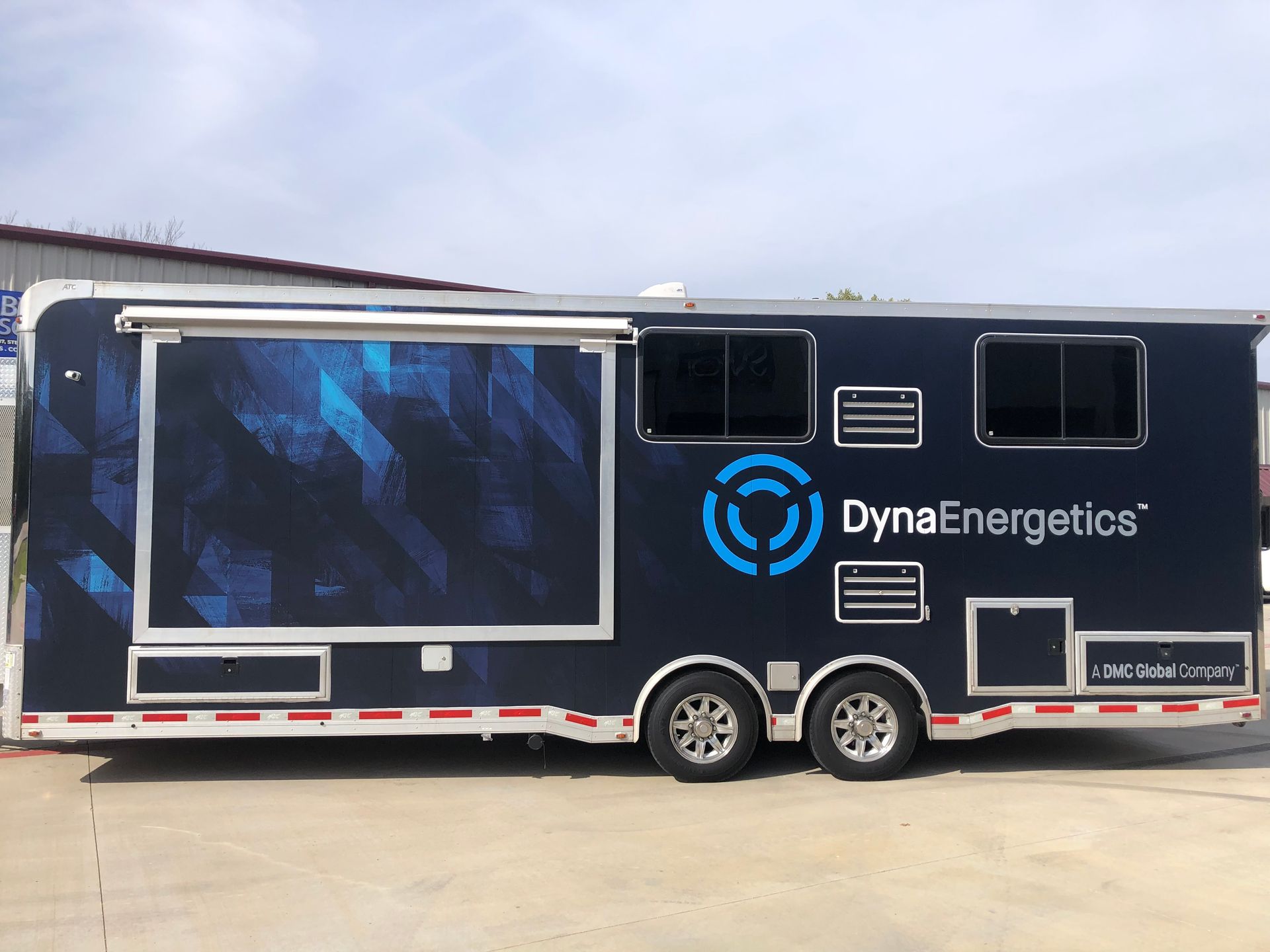 Black DynaEnergetics branded trailer with blue graphics, parked outdoors under a cloudy sky.
